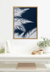 Framed cyanotype art of white palm leaves on a deep blue background, hanging above a white shelf with plants and books in a minimal room setting.