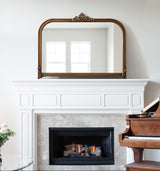 Arched wall mirror with ornate bronze frame above white traditional fireplace with lit fire and dark wood piano to the right in a bright room.