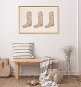 Framed artwork of three beige cowboy boots with subtle decorative stitching hanging above a wooden bench with striped cushion and throw in a minimalist room.