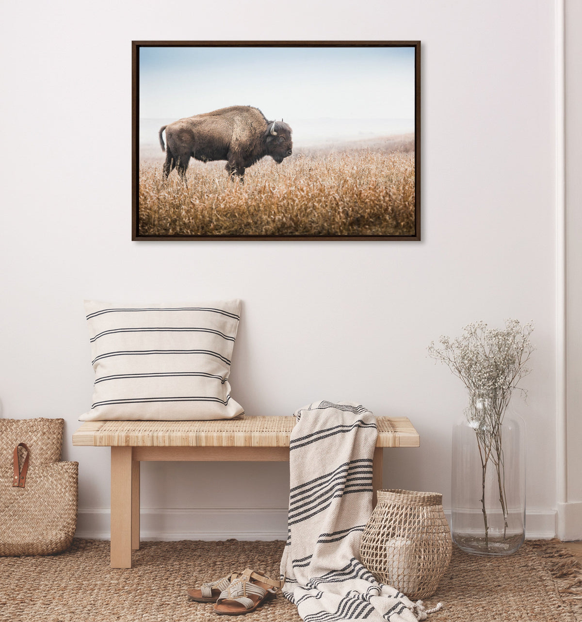 Framed canvas photo of a lone American bison standing in golden prairie grass under a pale blue sky, displayed above a wooden bench with striped textiles.