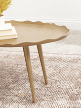Oval coffee table top with an irregular wavy edge in a matte gold finish, supported by slender tapered gold legs on a textured beige and brown rug.