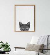 Framed canvas with a minimalist black kitten's face peeking from bottom, displayed above a white wall with a modern chair, neutral pillows, and green potted plant.