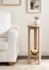 Tall, round wooden side table with three vertical legs and a lower shelf holding a gold and white decorative bowl, placed next to a white upholstered sofa.
