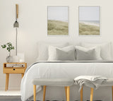 Minimalist bedroom with white bedding, gray pillows, a light wooden nightstand, and two framed coastal landscape artworks above the bed.