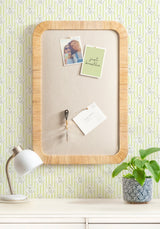 Rectangular pinboard with rounded wooden frame and beige fabric surface, displaying photos and notes, mounted on wallpaper with green and white floral pattern.