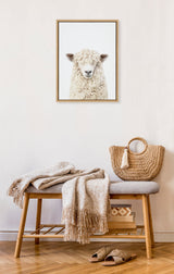 Framed portrait of a white sheep with fluffy wool and bangs on a plain light background, hanging above a bench with beige blankets, a woven bag, and slippers.