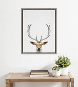 Framed canvas print featuring a detailed deer head with large antlers against a neutral background, hung above a wooden console with books and potted plants.
