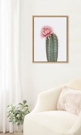 Framed artwork of a green cactus with a large pink flower on top, displayed on a white wall above a white armchair with a blush pillow and nearby plant.