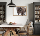Framed canvas of a gray and brown bison facing left on a white background, hung above a wooden dining table with mismatched chairs and gray cabinets.
