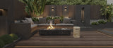 Outdoor seating area with gray sectional sofa, black rectangular fire pit, and textured round travertine marble side table on a dark wooden deck surrounded by greenery.