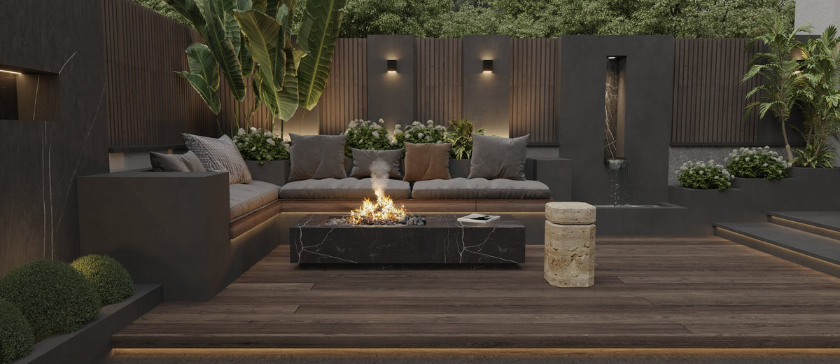 Outdoor seating area with gray sectional sofa, black rectangular fire pit, and textured round travertine marble side table on a dark wooden deck surrounded by greenery.