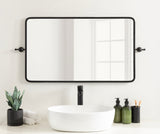 Rectangular wall mirror with thin black metal frame and rounded corners above a white oval sink, flanked by green glass dispensers and a small plant in a black pot