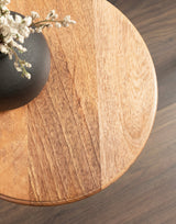 Close-up of a round wooden table top with visible grain patterns and a black vase holding small white flowers on the edge, set against a wood-paneled floor.