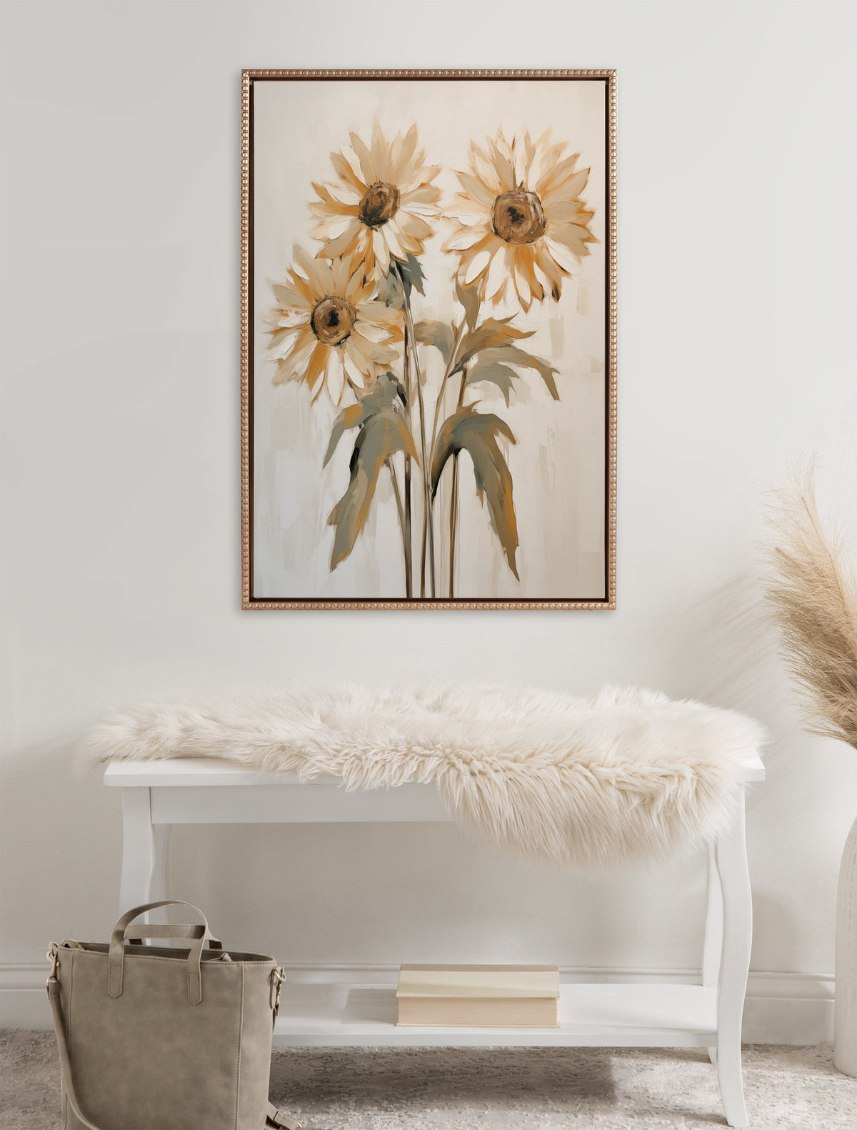 Framed canvas painting of three soft yellow sunflowers with green leaves on a light background, hanging above a white bench with a beige fur throw and taupe bag.