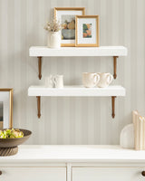Two white floating shelves mounted on a beige striped wall with bronze brackets; decorated with framed pictures, a vase with dried flowers, ceramic mugs, and a white pitcher.