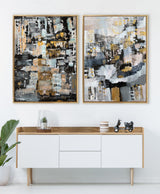 Two framed abstract paintings with black, gray, beige, and gold tones hang above a white and wood sideboard decorated with a glass vase, striped cups, and a small black scooter model in a minimalist room.