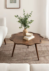 Dark brown scalloped wood coffee table with tapered legs, holding a beige vase with green leafy branches and two stacked off-white books topped by a small bowl, on a textured beige rug