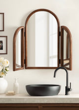 Arched tri-fold mirror with warm brown wooden frame above a concrete countertop holding a matte black vessel sink and matching matte black faucet.