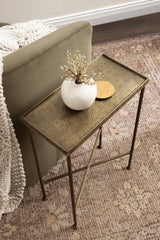 Slim metal side table with a textured bronze top and slender legs, holding a white round vase with dried flowers and a small gold dish, placed next to an olive sofa.