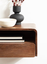 Close-up of a floating wooden side table with smooth rounded edges, featuring a black matte vase with dried flowers and a white ceramic bowl on top.