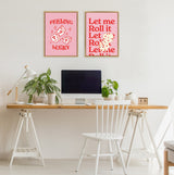 Two framed pink canvases with red text and dice illustrations hang above a minimalist wooden desk with white sawhorse legs, decorated with green plants and a white chair.
