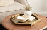 Brushed brass hexagonal tray with raised edges and rectangular handles sits on round wooden table, holding a white vase with small branches and a matching ceramic bowl.
