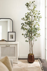 Tall artificial eucalyptus tree with round green leaves in a copper-colored pot beside a white wicker cabinet and arched mirror against a white wall.