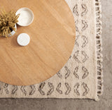 Top view of a round wooden coffee table on a textured beige rug with a brown pattern and tasseled edges, featuring a small white tray with dried flowers and candles.