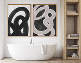 Two abstract black and white paintings with bold intertwining curved shapes hang above a white freestanding bathtub with a towel draped over its edge, next to a light wood shelving unit.