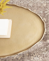 Oval coffee table with textured beige surface and slightly irregular wavy edges placed on a woven multicolored rug with a yellow floral accent and books on one corner.