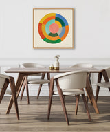 Square canvas with concentric circular segments in cream, yellow, orange, red, green, blue, and pink, framed in light wood and hung on a white wall above a wood dining table with cream upholstered chairs.
