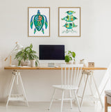 Minimalist home office with a wooden desk and white chair beneath two framed prints featuring stylized aquatic turtle and geometric family illustrations in green and blue tones.