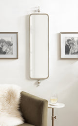 Tall, narrow rectangular mirror with rounded corners and a gray metal frame, mounted on a wall between two black-and-white framed photographs with decorative metal knobs above and below.