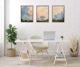Triptych of framed canvas paintings showing cloudscapes with blue, peach, and gray tones above green horizon, hung side by side on a white wall above a minimalist desk and chair.