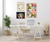 Three floral-themed framed canvases above a minimalist white desk with a laptop and chair, flanked by potted plants in woven baskets on light wood flooring.