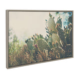 Framed canvas depicting a sunlit prickly pear cactus with green pads and brown spines surrounded by dry grass and blurred vegetation in the background.