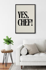 Black text 'YES, CHEF!' in bold serif font on a cream canvas framed in black, hung above a white cushioned sofa with neutral pillows and a wooden plant stand with a leafy green plant.