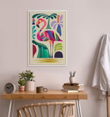 Colorful mid-century modern style flamingo artwork with bold pink, purple, green, and teal shapes in a white frame on a soft pink wall above a wooden desk with plants, books, and a chair.
