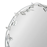 Round metal wall mirror with a delicate floral and butterfly design embellishing the upper left edge of the frame in a silver finish.