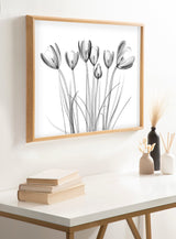 Black and white x-ray style floral artwork of crocus flowers with slender stems in a light wood frame, hanging above a white table with books and vases.