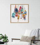 Watercolor painting of colorful vintage buoys hanging by ropes, framed in light wood on a white wall above a white chair with neutral cushions and a gray throw.