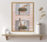 Framed artwork depicting two giraffes peeking from window planters with green foliage and pink flowers against pale peach building with grey shutters, hung above a wooden shelf with white vases.