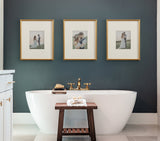 Three gold-beaded photo frames each with wedding couples portraits above a white freestanding bathtub against a dark gray wall, with a wooden stool holding folded towels and soap in front.