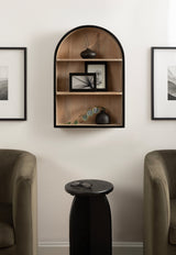Arched wood wall shelf with black trim holding a tall black vase with dried stems, two small black vases, photo frames, and a green branch, flanked by olive armchairs and black round side table.