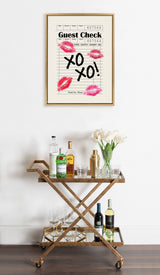 Framed canvas print of a guest check with red lipstick marks and black handwritten 'XO XO!' hanging above a wooden bar cart stocked with liquor bottles, glasses, and white tulips.