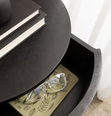 Partially opened dark gray curved drawer revealing a glasses case with leaf patterns and clear-framed glasses inside, beneath a round dark wood surface with stacked books.