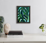 Framed 8x10 canvas painting of large green monstera leaves with dark and light green shades on a white wall above a white desk with books and plants.