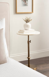Round white marble adjustable table with brass base and stem beside a bed, holding a white vase with dried flowers and stacked books, against a white wall with a framed photo.