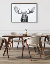 Black and white moose head portrait on a framed canvas hanging above a wood dining table with four cream-colored upholstered chairs in a minimalist room.