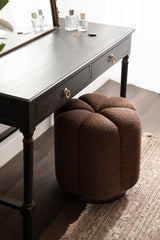 Brown round tufted ottoman with textured fabric placed under a dark wood desk with two drawers and brass ring pulls, on a patterned rug and wood floor.